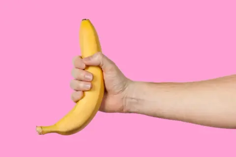Dłoń trzyma banana na różowym tle. Facet robi sobie dobrze, używając owocu.