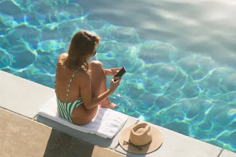 Kobieta w zielono-białym stroju kąpielowym siedzi przy basenie, trzymając telefon. Nie rób tego, kiedy on nie odpisuje, bo słońce i woda kuszą.