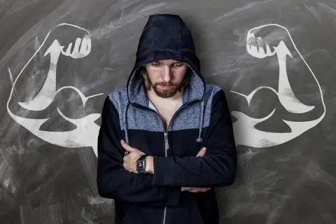 Chłopak w bluzie z kapturem, z ramionami skrzyżowanymi na piersi, stoi przed tablicą z rysunkami bicepsów. Idealny materiał na komplementy dla faceta, który czuje się silny.