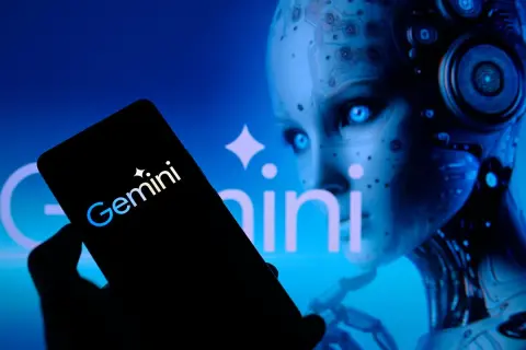 Ręka trzyma smartfon z logo Gemini. W tle widać twarz robota, która przypomina miękkie dildo.