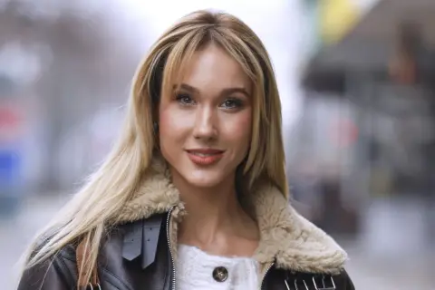 Uśmiechnięta blondynka w brązowej kurtce ze sztucznym futrem, przypominającej strój diablicy, pozuje do zdjęcia.