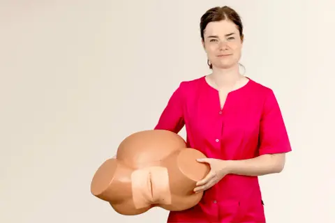 Kobieta w różowej bluzce trzyma model miednicy, prezentując anatomię do nauki masażu warg sromowych.